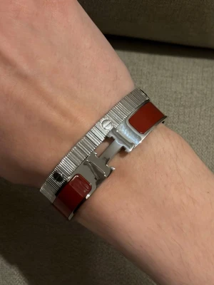 H armband i röd och silver - Snyggt H armband i silverfärgad metall med röda emaljdetaljer. Armbandet har H-formad lås och en stilren, modern design som passar perfekt till din outfit. Metall och emalj ger en lyxig känsla.