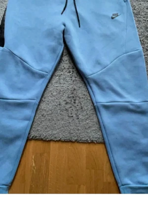 Ljusblå Nike mjukisbyxor - Snygga ljusblå mjukisbyxor från Nike med svarta detaljer och logga på vänster höft. Byxorna har resår i midjan med dragsko, ribbade muddar vid bensluten och en svart dragkedja på ena fickan. Perfekta för en avslappnad och sportig stil.