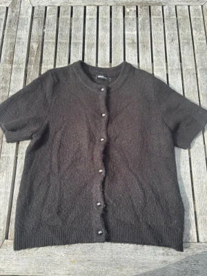 Svart kortärmad kofta Gina Tricot - Kortärmad svart kofta från Gina Tricot med rund halsringning och dekorativa pärlknappar framtill. Koftan är stickad i ett mjukt material och har ribbade muddar vid ärmslut och nederkant. På bilden skiftar färgen i brun och svart på grund av solen, men i verkligheten är den endast svart👍🏻