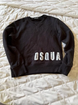 Svart sweatshirt från Dsquared2 - Cool svart sweatshirt från Dsquared2 med stor vit DSQUA-text framtill. Tröjan har rund halsringning, ribbade muddar vid ärmslut och nederkant samt mjukt, borstat innerfoder för extra komfort. Perfekt för en avslappnad och trendig look.