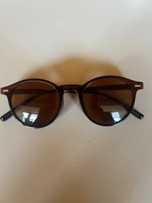 Runda bruna solglasögon - Snygga runda solglasögon med bruna bågar och matchande bruna glas. Klassisk retrostil som passar perfekt till sommaren. Bågarna är i plast och har en tidlös look som funkar till många outfits.