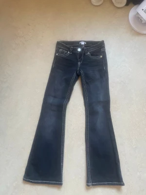Svarta bootcut jeans med kontrastsömmar - Snygga svarta bootcut jeans med vita kontrastsömmar och klassiska fem fickor. Jeansen har låg midja och utsvängda ben, samt dekorativa sömmar på bakfickorna. Perfekta för en trendig och avslappnad stil. Använd 1 gång
