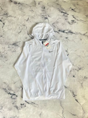 Vit hoodie med dragkedja från Nike - Snygg vit hoodie från Nike med dragkedja framtill och diskret Nike-logga på bröstet. Tillverkad i ett lätt och sportigt material, perfekt för en avslappnad stil. Huva och långa ärmar med resår vid handlederna ger en clean look.