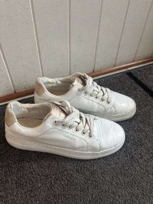 Vita sneakers från Gant - Stilrena vita sneakers från Gant med lågt skaft och klassisk design. Skorna har vita snören, diskret Gant-logga på sidan och detaljer i beige vid hälen. Ovandelen är i slätt läder och sulan är vit med ljusbrun undersida.
