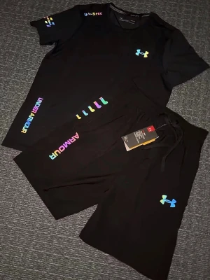 Svart träningsset från Under Armour - Säljer ett par svart träningsset från Under Armour med färgglada loggor och textdetaljer. Shortsen har snörning i midjan och är perfekta för träning eller chill. Matchar med t-shirt i samma stil för en komplett look. Storlek S, helt ny