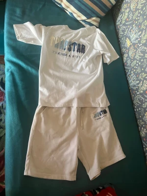 Vitt Trapstar set med tryck - Säljer ett vitt set från Trapstar med matchande t-shirt och shorts. Båda delarna har trycket 'Trapstar It's a Secret' i blå och grå toner på bröstet och benet. Setet är gjort i mjukt bomullsmaterial och har en avslappnad passform, perfekt för chill dagar.