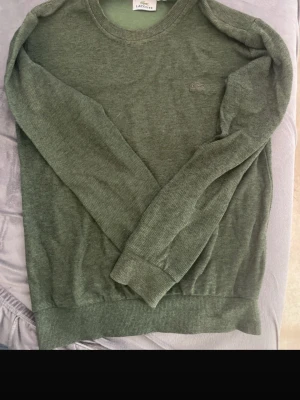 Grön stickad tröja från Lacoste - Mysig grön stickad tröja från Lacoste med rund halsringning och diskret krokodillogga på bröstet. Tröjan har långa ärmar och ribbade muddar vid ärmslut och nederkant. Perfekt för lager på lager och enkel att matcha.