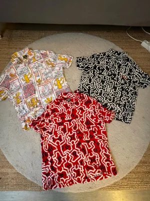 Färgglada skjortor med grafiskt mönster - Tre skjortor med ikoniska Keith Haring-motiv i vitt, svart och rött. Alla har kort ärm, knappar framtill och ett unikt grafiskt mönster. Perfekt för dig som gillar streetstyle och konstnärliga prints.
