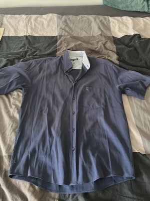Blå kortärmad skjorta Tommy Hilfiger - Snygg blå skjorta från Tommy Hilfiger med korta ärmar och klassisk krage. Skjortan har knappar framtill och en bröstficka med liten logga. Perfekt för en stilren och avslappnad look.