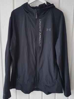 Svart träningsjacka från Under Armour - Snygg svart träningsjacka från Under Armour med hel dragkedja och huva. Jackan har logga och text på bröstet, samt två sidofickor med dragkedja. Tillverkad i lätt och funktionellt material som passar perfekt till träning och vardagsaktiviteter.