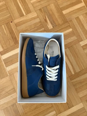 Blå Maison Margiela gats storlek 43 - Snygga blå sneakers från Maison Margiela med vita skosnören och beige gummisula. Skorna har en mix av mocka och läderpaneler för en stilren look. Klassisk låg modell som passar perfekt till jeans eller streetwear.