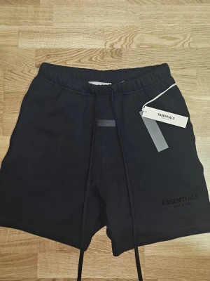 Svarta shorts från Essentials Fear of God - Säljer ett par svarta shorts från Essentials Fear of God med elastisk midja och långa snören framtill. Shortsen har en clean look med diskret logga på benet och är perfekta för en avslappnad stil. Tillverkade i mjukt material för maximal komfort.