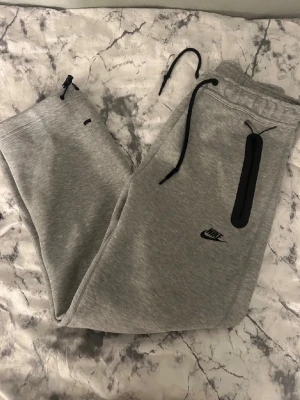 Grå Nike mjukisbyxor med svarta detaljer - Snygga grå mjukisbyxor från Nike med svarta dragsnören och en svart dragkedja vid fickan. Byxorna har en smal passform och Nike-logga på låret. Perfekta för en avslappnad och sportig stil.