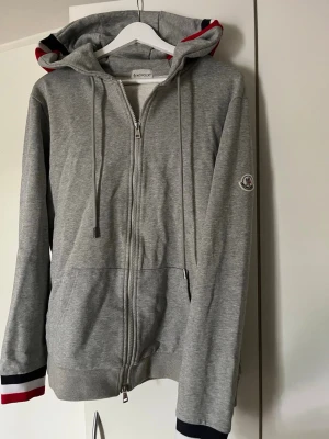 Grå zip hoodie från Moncler - Snygg grå hoodie från Moncler med dragkedja, huva med dragsko och Moncler-logga på ärmen. Ribbade muddar med röd, vit och marinblå detalj samt matchande färger i luvan. Perfekt för en chill och stilren look.