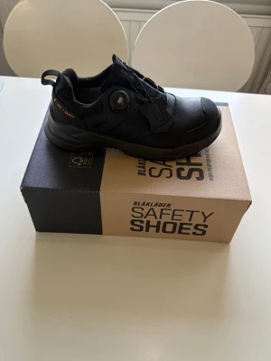 Svarta sneakers från Blåkläder - Svarta sneakers från Blåkläder med robust design och smart snörningssystem. Skorna har detaljer i svart och orange, samt en slitstark ovandel i läder och textil. Perfekta för dig som vill ha en cool och funktionell look.
