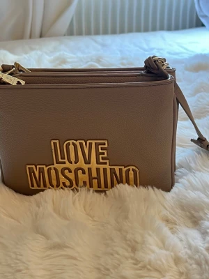 Beige axelväska från Love Moschino - Snygg beige axelväska från Love Moschino i mjukt konstläder. Väskan har två stora fack med guldfärgade dragkedjor och ett stort guldfärgat logotypmärke framtill. Justerbar axelrem och detaljer i guld ger en lyxig känsla.