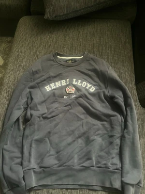 Grå sweatshirt från Henri Lloyd - Mjuk grå sweatshirt från Henri Lloyd med tryckt logga och text 'Est. 1963' på bröstet. Rund halsringning och långa ärmar. Perfekt för en avslappnad och stilren look.