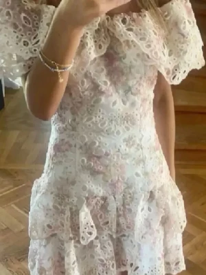 Vit spetsklänning med volanger - Superfin populär kort vit klänning med broderad spets, offshoulder och volangdetaljer. Klänningen har ett blommigt mönster i ljusa toner och flera lager för extra volym. Perfekt för sommarens alla tillfällen!