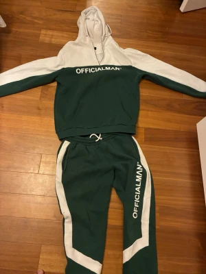 Grön och vit -set OFFICIALMAN - Tvådelat set från OFFICIALMAN i grönt och vitt. Setet består av en hoodie med huva och matchande byxor med dragsko i midjan. Båda delarna har vita detaljer och logotypen tryckt i vitt. Perfekt för chill dagar eller när du vill vara bekväm.