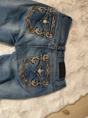 Blå jeans Shyanni  - Säljer ett par blå jeans med snygga broderade detaljer i beige på bakfickorna och knappar på ficklocken. Jeansen är bootcut och har en  klassisk femficksdesign. Perfekta för dig som vill ha något unikt och stilrent i garderoben. Pris kan diskuteras 