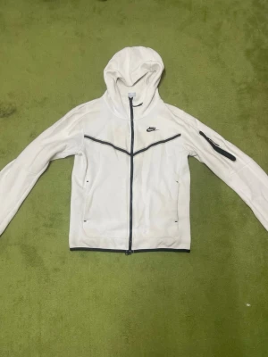 Vit hoodie från Nike med dragkedja - Snygg vit hoodie från Nike med hel dragkedja framtill och svarta detaljer. Hoodien har långa ärmar, fickor på sidorna och en extra ficka med dragkedja på ena ärmen. Perfekt för en sportig och avslappnad stil.