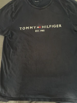 Svart Tommy Hilfiger t-shirt - Klassisk svart t-shirt från Tommy Hilfiger med tryckt logga och texten 'EST. 1985' på bröstet. Rund halsringning och korta ärmar. Perfekt för en enkel och stilren look.