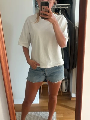 Blå jeansshorts med fransig kant - Säljer ett par klassiska blå jeansshorts med rå, fransig kant och femficksmodell. Shortsen har normal passform och är tillverkade i bomull. Perfekta för sommaren och enkla att matcha med olika toppar.