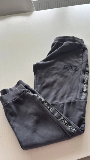 Gråa mjukisbyxor från EA7 Armani - Snygga grå mjukisbyxor från EA7 Emporio Armani med logga på benet och svarta sidoband med vit text. Byxorna har resår i midjan och är gjorda i mjukt bomullsmaterial. Perfekta för chill dagar eller när du vill ha en sportig look.
