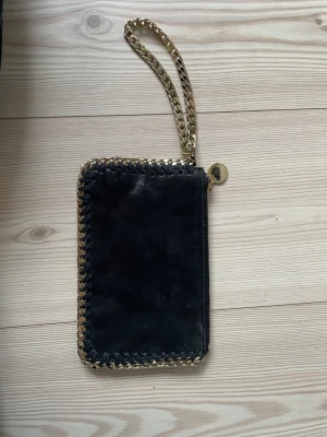 Stella McCartney plånbok  - Snygg svart plånbok från Stella McCartney med guldfärgad kedja runt kanterna och som handledsrem. Plånboken har dragkedja och en rund guldfärgad logga. Materialet är veganskt mockaliknande tyg.
