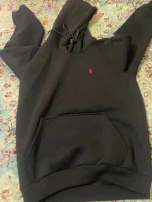 Svart hoodie från Polo Ralph Lauren - Snygg svart hoodie från Polo Ralph Lauren med klassisk röd logga broderad på bröstet. Hoodien har huva med dragsko och en stor magficka framtill. Tillverkad i mjukt material som känns skönt mot huden. Perfekt för en avslappnad och stilren look.