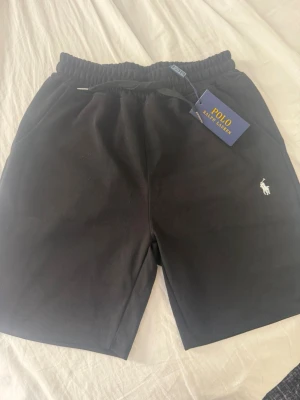 Svarta shorts från Polo Ralph Lauren - Svarta shorts från Polo Ralph Lauren med klassisk vit logga broderad på vänster ben. Shortsen har elastisk midja med snörning och är tillverkade i mjuk bomull, perfekta för en avslappnad och stilren look.