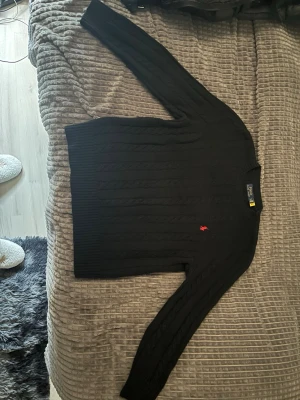 Svart kabelstickad tröja Polo Ralph Lauren - Kabelstickad svart tröja från Polo Ralph Lauren med rund halsringning och lång ärm. Tröjan har den klassiska röda logotypen broderad på bröstet och ribbade muddar vid ärmslut och nederkant. Perfekt för en stilren och tidlös look.