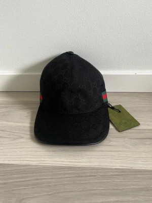Svart keps från Guc ci med GG-mönster - Snygg svart keps från Guc ci med klassiskt GG-monogram i tyget och gröna samt röda detaljer på sidan. Kepsen har böjd skärm och justerbar rem bak. Tillverkad i slitstarkt tyg och levereras med originaltagg.