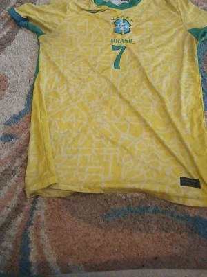 Brasilien Vini Jr. matchtröja gul - Säljer en gul Brasilien matchtröja med gröna detaljer och nummer 7, VINI JR. på ryggen. Tröjan har ett grafiskt mönster, rund hals och korta ärmar. Perfekt för dig som älskar fotboll och vill stötta landslaget.
