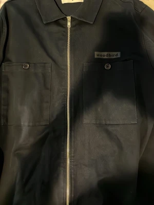 Svart overshirt från Woodbird - Stilren svart overshirt från Woodbird med två stora bröstfickor med knapp, rak passform och dragkedja framtill. Overshirten har klassisk krage och diskret logga på bröstet. Perfekt lager-på-lager-plagg för en modern look.