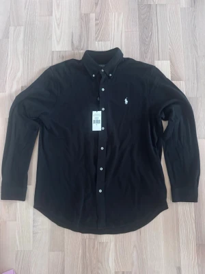 Svart skjorta från Ralph Lauren - Klassisk svart skjorta från Ralph Lauren med button-down krage och vita knappar. Skjortan har lång ärm och den ikoniska logotypen broderad på bröstet. Tillverkad i mjuk bomull för en bekväm känsla och snygg passform.