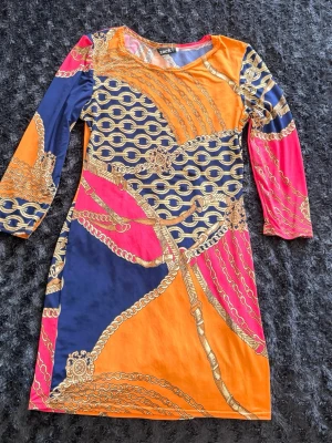 Färgstark mönstrad klänning från Zack - Cool klänning från Zack med färgstarkt kedjemönster i orange, marinblått, rosa och gult. Modellen är kort med trekvartsärm och rund halsringning. Perfekt för dig som gillar att sticka ut med unika prints och färger.