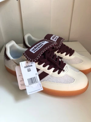 Adidas Wales Bonner sneakers vita/vinröda - Snygga Adidas Wales Bonner sneakers i vitt och vinrött med klassiska tre ränder på sidan. Skorna har en unik tungdesign med kontrastsömmar, vinröda skosnören och gummisula. Materialmix av läder och textil ger en retrovibe och cool look.