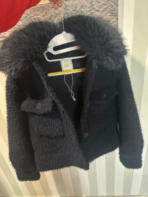 Svart teddyjacka med fuskpäls - Fin marinblå teddyjacka med stor fluffig fuskpäls-krage. Jackan har två bröstfickor med knappar och stängs med fyra stora knappar framtill. Kortare modell som ger en trendig look. Perfekt för dig som vill ha en mysig och stilren jacka till hösten eller vintern. Använd bara 2-3 gånger mycket bra skick, pris går att diskutera köpt på Gina tricot för 400kr tror jag 