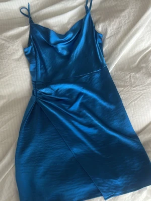 Blå satin klänning med smala band - Snygg blå klänning i satin med smala axelband och omlott-detalj framtill. Klänningen har en lätt glansig finish och figurnära passform som ger en elegant look. Perfekt för dig som vill sticka ut med en färgstark och trendig stil.