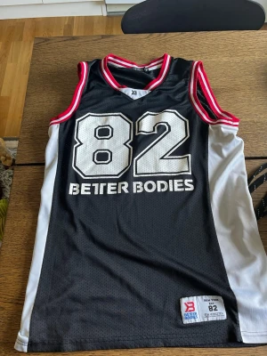 Basketlinne från Better Bodies - Snyggt basketlinne från Better Bodies i svart med vita sidopaneler och stora siffran 82 framtill. Röd och vit randig kant vid hals och ärmhål. Tillverkat i meshmaterial som andas, perfekt för träning eller sport.