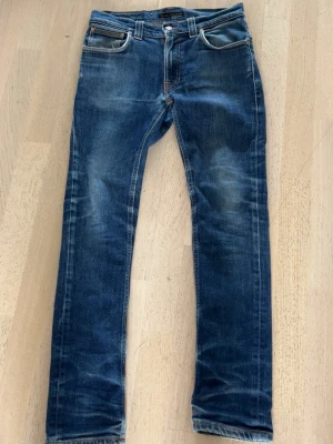 Blå slim fit jeans från Nudie - Snygga blå slim fit jeans från Nudie med klassisk femficksdesign och slitna detaljer. Jeansen har orange sömmar och broderade bakfickor. Tillverkade i bomull med en skön stretch för extra komfort.