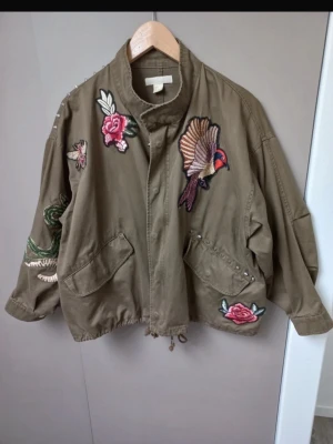 Grön jacka med coola mönster - Cool grön parkajacka med broderade patchar av rosor, fågel och orm. Jackan har dragsko i midjan, stora 