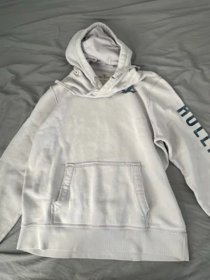 hoodie från Hollister - Snygg rosa hoodie från Hollister med stor känguruficka framtill och blå logga på ryggen. Ärmen har texten 'HOLLISTER' tryckt i blått. Klassisk huva och mjukt material som känns skönt mot huden. Perfekt för en avslappnad och cool stil.
