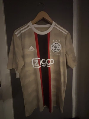 Ajax fotbollströja Adidas - Ajax tredje ställ från 2022/2023 säsongen. Riktigt snygg beige tröja med svarta och röda ränder längs mitten. Stilrena vita detaljer och även bilder på arenor samt kända byggnader från Amsterdam. Gjord i bekvämt och luftigt material perfekt för att röra sig i eller bara ha som en skön tröja under sommaren. 176 i storlek så skulle säga att det är runt S eller M. Inga skador, fläckar eller missfärgningar. Priset är inte hugget i sten så kan tänka mig att gå ned lite.