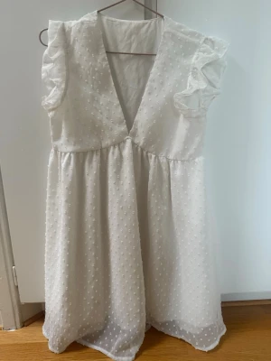 Studentklänning  - Söt vit kortklänning med v-ringning och volangärmar. Klänningen har ett prickigt mönster i tyget och är lätt och luftig. Den har en lös passform och är ärmlös med volangdetaljer. Förekommer en liten sminkfläck, se bild. 