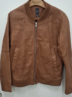 Brun skinnjacka från Emilio - Snygg brun skinnjacka från Em7lio med ribbade muddar vid krage, ärmslut och nederkant. Jackan har dragkedja framtill, två sidofickor och en stilren, minimalistisk design. Perfekt för dig som gillar klassisk men modern look.