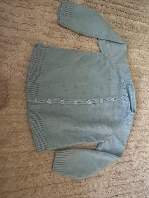 Ljusblå kofta med prickiga knappar - Söt ljusblå kofta i mjukt stickat material med långa ärmar och ribbade muddar. Framtill sitter dekorativa blommiga knappar som ger en gullig touch. Perfekt att slänga över en topp för en avslappnad look. 