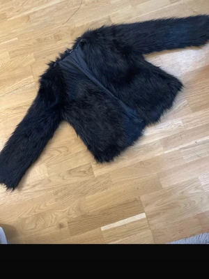 Svart fuskpälsjacka med lång lugg - Snygg svart fuskpälsjacka med lång lugg och helsvart innerfoder. Jackan har rak passform och långa ärmar, perfekt för att hålla värmen under kalla dagar. Stängs framtill och ger en cool look till din outfit.