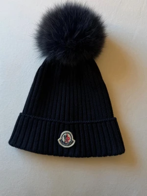Svart Moncler mössa med pälsboll - Snygg svart ribbstickad mössa från Moncler med stor fluffig pälsboll på toppen och klassisk Moncler-logga framtill. Perfekt accessoar för kalla dagar och ger en lyxig touch till din outfit. Köpt för 3975